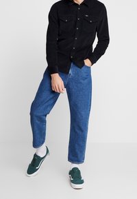 Czarna koszula z kordurowego materiału z dwiema kieszeniami na piersi, noszona z luźnymi niebieskimi dżinsami i zielonymi sneakersami. Białe skarpetki widoczne na kostkach.