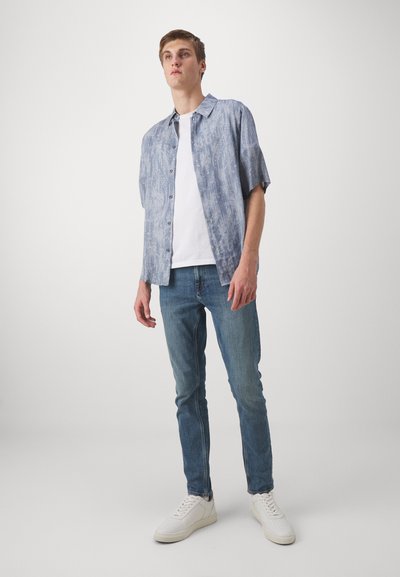 Chemise à manches courtes, de couleur bleu clair, à motifs, avec un col, portée par-dessus un T-shirt blanc. Associée à un jean bleu et des baskets blanches.