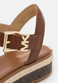 Sandale compensée en cuir marron avec boucle dorée et logo "MK" sur la bride, bordure tissée beige, et semelle plateforme foncée à motifs.