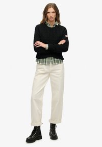 Zwarte kabelgebreide trui over een groen-wit geruit overhemd, gecombineerd met roomkleurige broek met wijde pijpen en zwarte veterlaarzen.