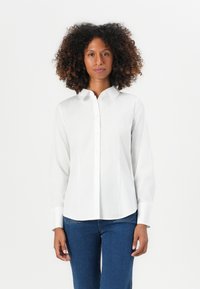 Donna con capelli ricci che indossa una camicia bianca con bottoni e jeans blu, in piedi contro uno sfondo bianco uniforme.