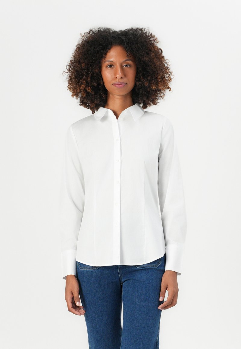 Donna con capelli ricci che indossa una camicia bianca con bottoni e jeans blu, in piedi contro uno sfondo bianco uniforme.
