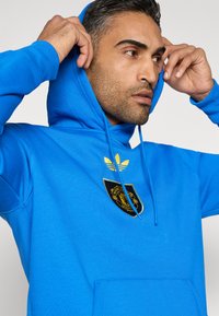 Modrá mikina s kapucňou z mäkkého materiálu, s čiernym logom Manchester United a zlatými prúžkami Adidas na hrudi. Textúrovaná s klokanou kapsou.