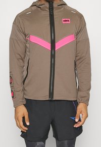 Nike-jas in bruin met een roze chevron-ontwerp. Voorzien van een rits aan de voorkant, capuchon en elastische manchetten. Logo op de borst en details op de mouwen.