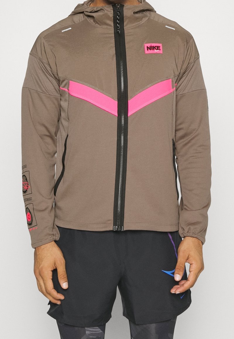 Nike-jas in bruin met een roze chevron-ontwerp. Voorzien van een rits aan de voorkant, capuchon en elastische manchetten. Logo op de borst en details op de mouwen.