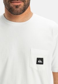 Bílý bavlněný tričko s náprsní kapsou, na které je černá nálepka se sloganem. Krátké rukávy a klasický kulatý výstřih.