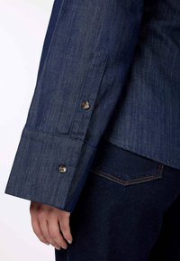 Manica di camicia in denim blu scuro con polsino a doppio bottone e una texture liscia con dettagli di cucitura sottili.