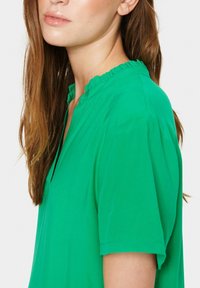 Haut vert à manches courtes avec un col volanté, offrant une coupe décontractée et un tissu doux. L'encolure a une forme en V subtile.