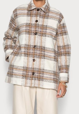 Vrouw die een beige, bruin en grijs geruit jasje met knopen en lange mouwen draagt, gecombineerd met crèmekleurige wijde broek.