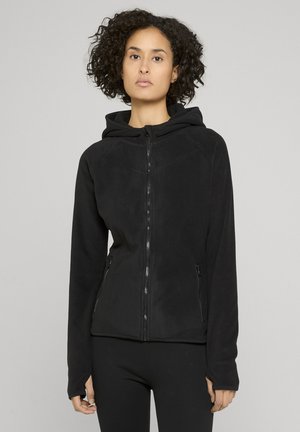 Donna con capelli ricci che indossa una felpa nera con zip e tasche anteriori, in piedi davanti a uno sfondo grigio chiaro uniforme.