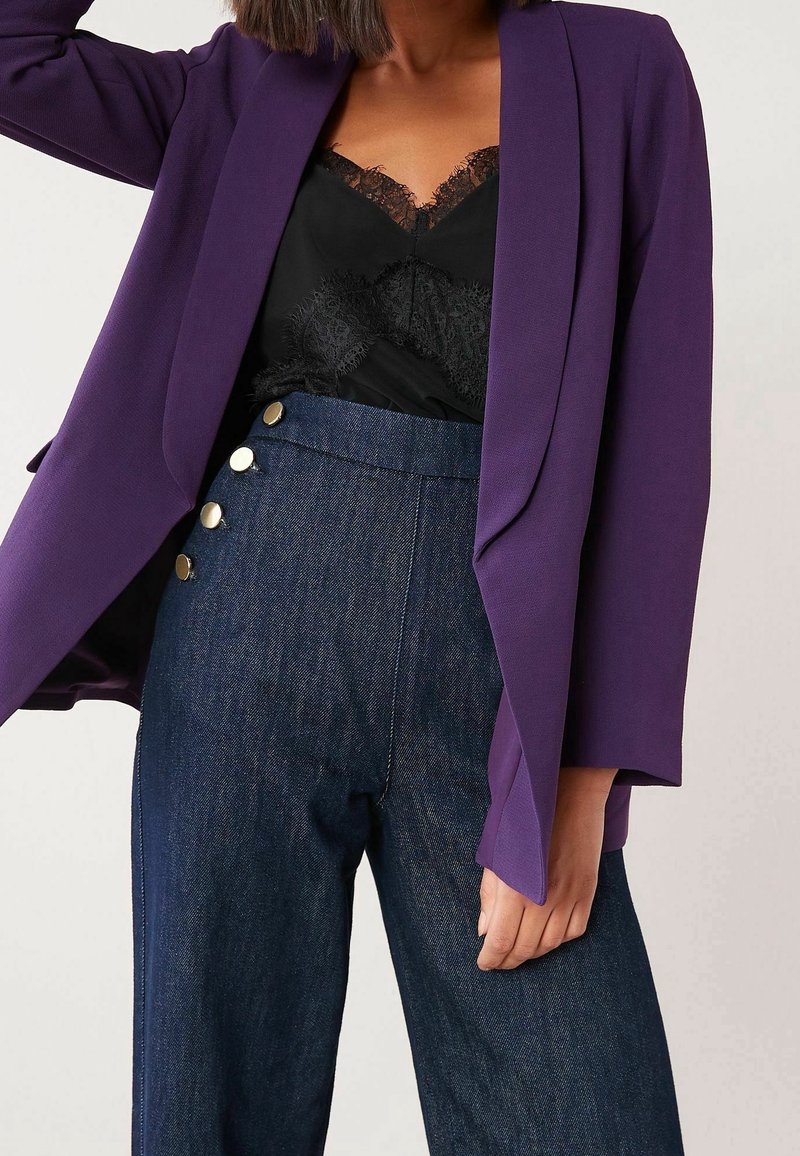 Blazer violet à coupe décontractée, associé à un caraco noir orné de dentelle et un pantalon en denim foncé taille haute avec des boutons dorés en accent.
