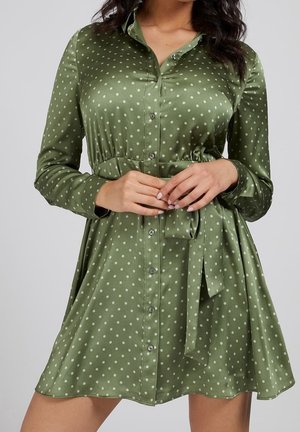 Femme portant une robe chemise en satin vert à pois, nouée à la taille, manches longues, et boutonnée devant, mains jointes à la taille.