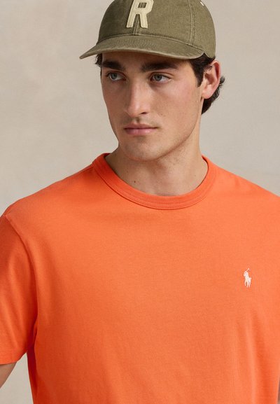 Fiatal férfi olívazöld sapkával, amin az "R" betű látható, és élénk narancssárga pólóval, fehér hímzett logóval, jobbra nézve.