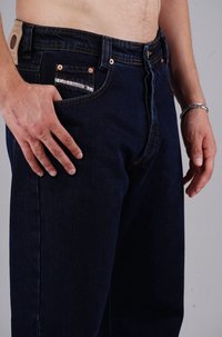 Dunkelblaue Jeans mit klassischer Passform, ausgestattet mit kupferfarbenen Beschlägen, kontrastierenden Nähten und einem Etikett am Bund. Fronttaschen-Detail sichtbar.