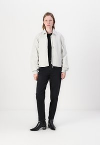 Helle graue Jacke mit Reißverschluss und elastischen Bündchen, in lockerer Passform, kombiniert mit schwarzen Hosen und Ankle Boots. Einfarbiger Hintergrund, keine Muster oder Akzente.
