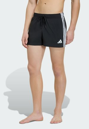 Mężczyzna w czarnych kąpielówkach Adidas z białymi bocznymi paskami i logo, stoi boso na jednolitym jasnym tle.