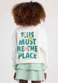 Felpa bianca con testo verde "QUESTO DEVE ESSERE IL POSTO" e vari adesivi grafici colorati. Design leggero e casual con una vestibilità comoda.