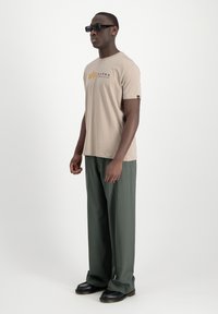 Camiseta de manga corta beige con el logo "ALPHA" en dorado. Combinada con pantalones anchos verdes. El modelo también lleva gafas de sol negras y zapatos.