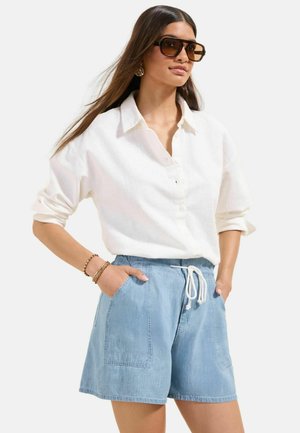Kvinde iført en stor hvid skjorte med knapper, lyse denimshorts med snøre, store firkantede solbriller og guldbøjler, hænderne i lommerne.