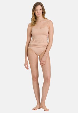Femme debout pieds nus portant une camisole couleur chair et des sous-vêtements assortis sur un fond clair uni.
