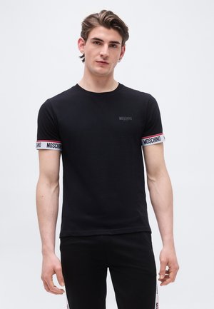 Jeune homme portant un t-shirt Moschino noir avec le logo sur les manches et la poitrine, debout devant un fond clair uni.