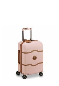Valise rigide de couleur rose pâle avec une surface texturée. Présente des accents marron, une poignée rétractable et quatre roues noires pour la mobilité.