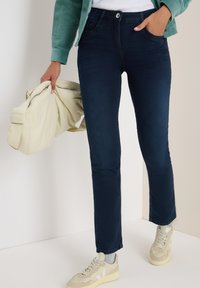 Vrouw in donkerblauwe jeans en beige sneakers, met een lichtbeige jas in één hand, staande tegen een neutrale achtergrond.