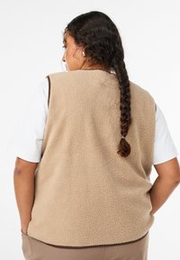 Beige fleecevest med en tekstureret overflade, med brune kanter langs kanten, båret over en hvid skjorte, med en synlig siden lomme.