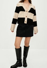 Black and cream striped cropped cardigan with a V-neck, button closure, and ribbed cuffs, paired with a black denim skirt and knee-high boots.