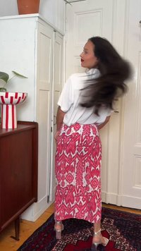 Γυναίκα με μακριά σκούρα μαλλιά, που φοράει λευκό πουκάμισο και ροζ φούστα με σχέδια, γυρίζει το κεφάλι της, στέκεται μέσα σε δωμάτιο με λευκές πόρτες και ξύλινα έπιπλα.