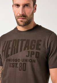 Hnědý bavlněný tričko s velkým výrazným potiskem "HERITAGE JPD INDIGO UNION EST.80." Měkká textura, kulatý výstřih, krátké rukávy.