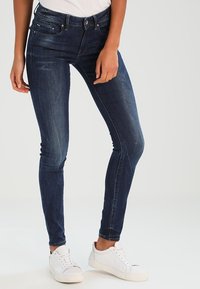 Vrouw in donkerblauwe skinny jeans en witte sneakers, staande met één hand rustend nabij de dij tegen een effen achtergrond.