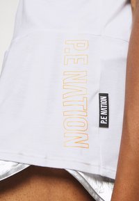 Débardeur blanc sans manches avec le texte "P.E NATION" en contour jaune et une étiquette en tissu noir portant "P.E NATION" en lettres blanches.