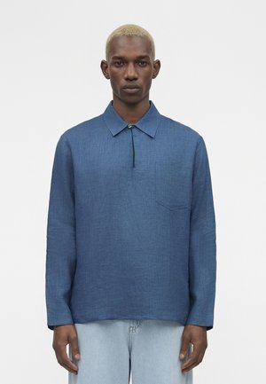 Muž s krátkymi blond vlasmi, oblečený v modrej košeli s dlhým rukávom a golierom a svetlomodrých džínsoch, stojaci pred obyčajným pozadím.