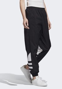 Svarta vindjacka-byxor med elastisk midja, avsmalnande ben och vita detaljer med randigt mönster. Bärs med vita sneakers.