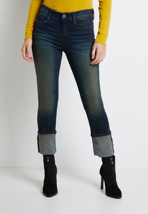 Jean skinny avec un lavage indigo foncé, un léger délavage, et un revers contrastant en denim plus clair. Comprend des poches standard et des fermetures à boutons en métal.
