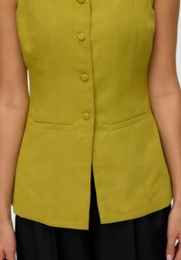 Gilet en lin vert sans manches, à coupe droite, avec six boutons ronds et deux poches avant. Jupe noire sous le gilet.