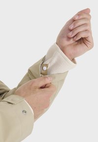 Beige jakkeærme med en hvid fleeceforing og trykknaplukning. Hånd der griber om ærmet, hvilket viser en glat tekstur og diskrete syninger.