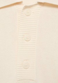 Kermasoitune polo-paita, jossa on ribbed-kenttä, kolme napin sulkemista ja sileä pinta. Napit ovat vaaleanväristä muovia.