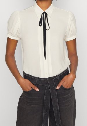 Persona que lleva una blusa blanca de manga corta con una corbata negra, combinada con unos vaqueros negros de talle alto con cinturón, con las manos en los bolsillos.