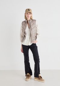 Gilet matelassé beige sur un pull blanc à détails de boutons, associé à un pantalon noir et des baskets marron clair avec des à-tassels ; portant une casquette beige.