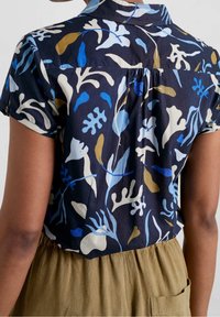 Bakvy av en person som bär en marinblå kortärmad skjorta med abstrakta vita, blåa och bruna bladmönster, insatt i beige byxor med ficka.