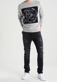 Hombre vestido con sudadera gris con estampado gráfico y vaqueros ajustados desgastados negros, con zapatillas blancas, de pie frente a un fondo blanco.