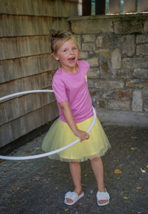 Lächendes Mädchen in pinkem Shirt und gelbem Tüllrock, das im Freien in der Nähe von Stein- und Holzwänden einen weißen Hula-Hoop-Reifen dreht.
