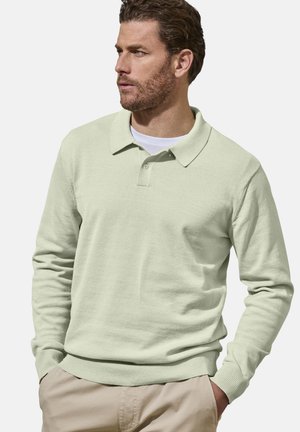 Mann mit Bart, der ein hellgrünes langärmeliges Poloshirt und beige Hose trägt, schaut nach links und hat die Hände in den Taschen vor einem einfarbigen Hintergrund.