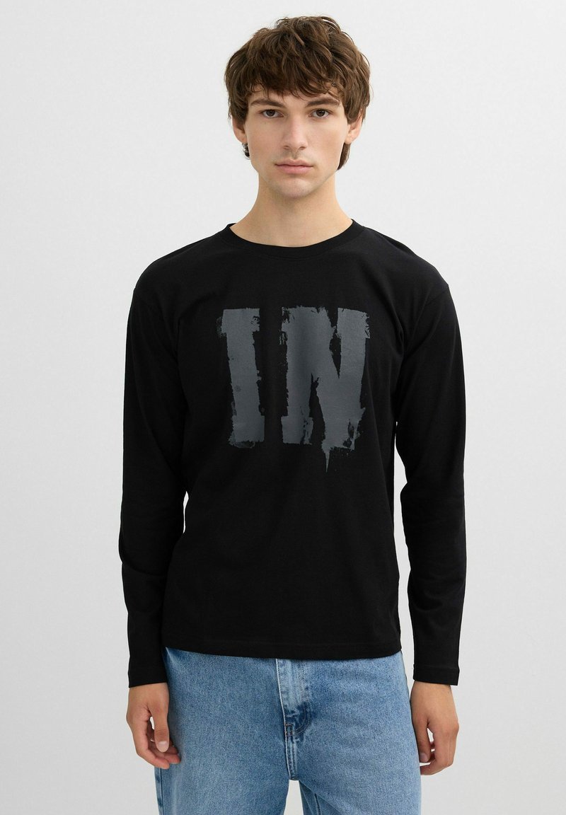 Camiseta negra de manga larga con una gran impresión en gris desgastado que dice "IN" en el centro. Material de algodón, corte casual, combinada con jeans de color azul claro.
