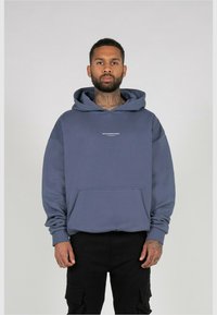 Blauwe katoenen hoodie met een voorzak en een verstelbare capuchon, met een witte opdruk van het logo op de borst. Model draagt zwarte broek.
