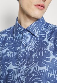 Kortärmad skjorta med knäppning i ljusblått med ett mörkblått mönster av tropiska blad. Den har krage, två blå knappar och en texturerad tyg.