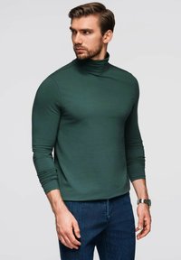 Dunkelgrüner Rollkragenpullover mit langen Ärmeln, enganpassendem Design, weicher Textur und minimalen Nähten, kombiniert mit blauen Jeans.