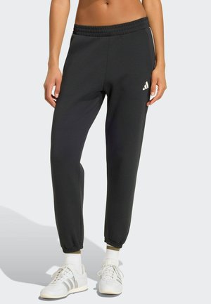 Kvinde iført sorte Adidas joggingbukser med elastiske manchetter og hvide sneakers, stående mod en ensfarvet lys baggrund.